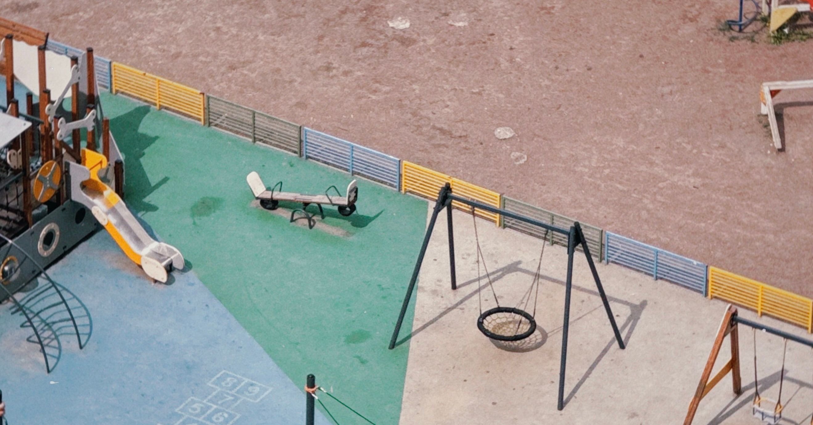 Aerial view of an empty outdoor playground with blue, green and beige surfacing, a slide, a seesaw and swings.