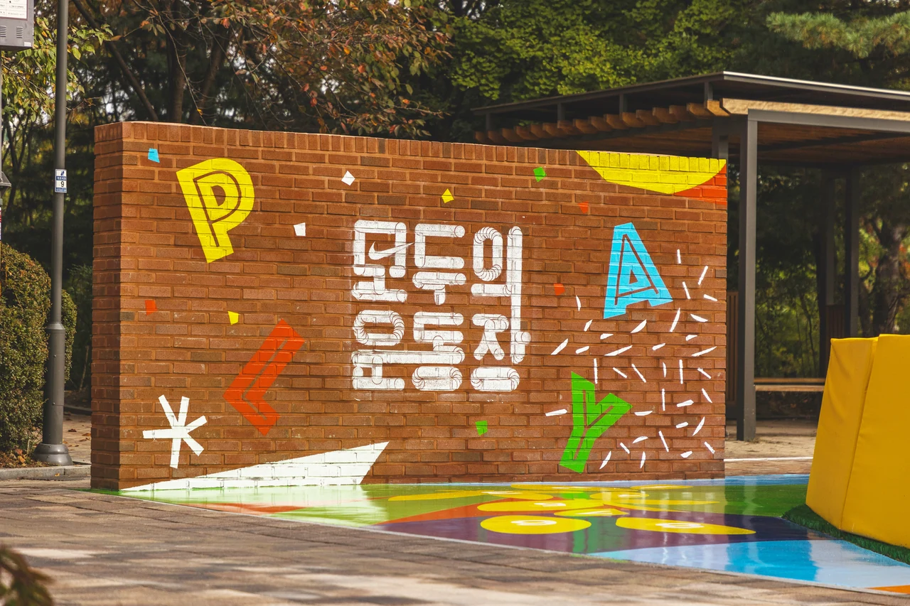 A red brick wall painted with the Modoo project lettering and graphics, beside a colourful ground surface.