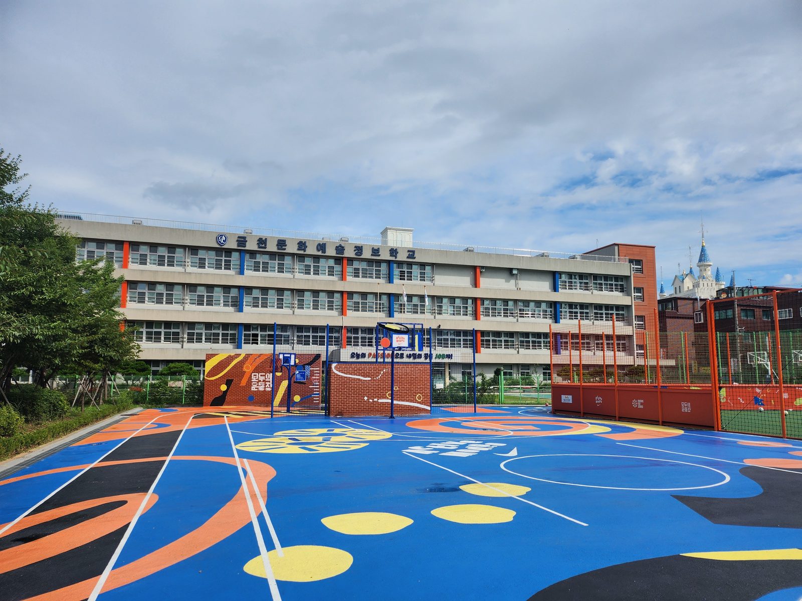 A blue multi-use court with yellow, orange and black graphics in front of a school building.